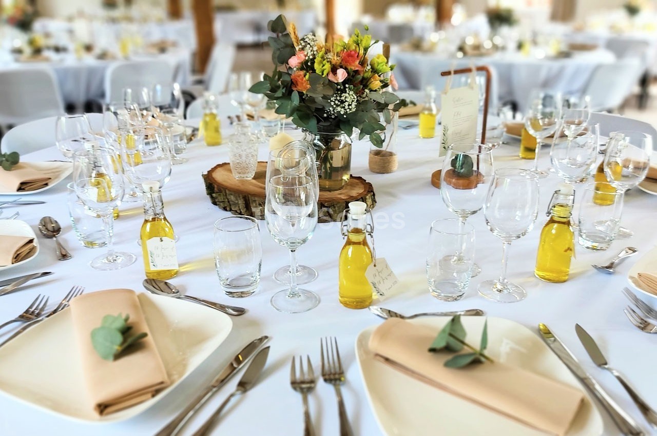Table dressée pour un repas festif avec nappes blanches, fleurs au centre, couverts, verres et petites bouteilles d'huile.
