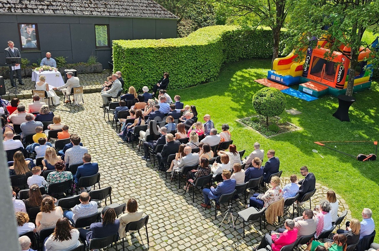 Cérémonie en plein air avec des invités assis, un officiant, une table décorée et une structure gonflable en arrière-plan.