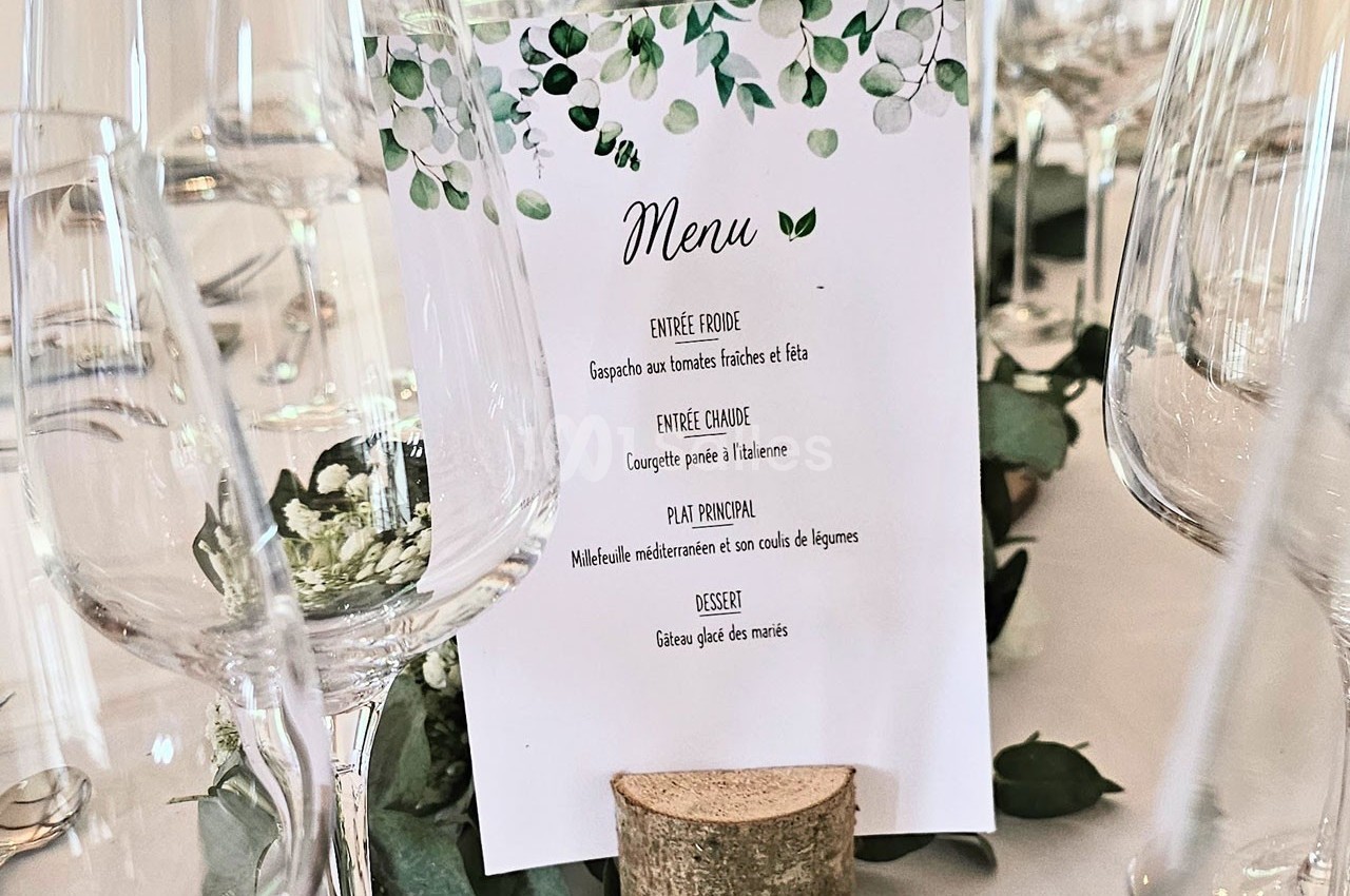Carte de menu imprimée sur papier blanc avec motifs végétaux, posée sur un rondin de bois entourée de verres.