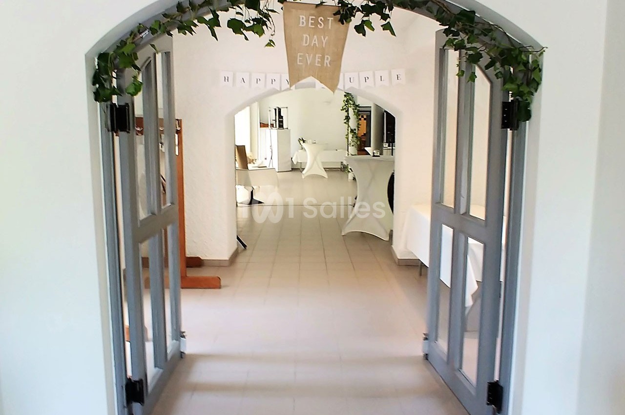 Entrée d'une salle décorée pour un événement, avec une arche ornée de feuillage et une bannière ’Best Day Ever’.