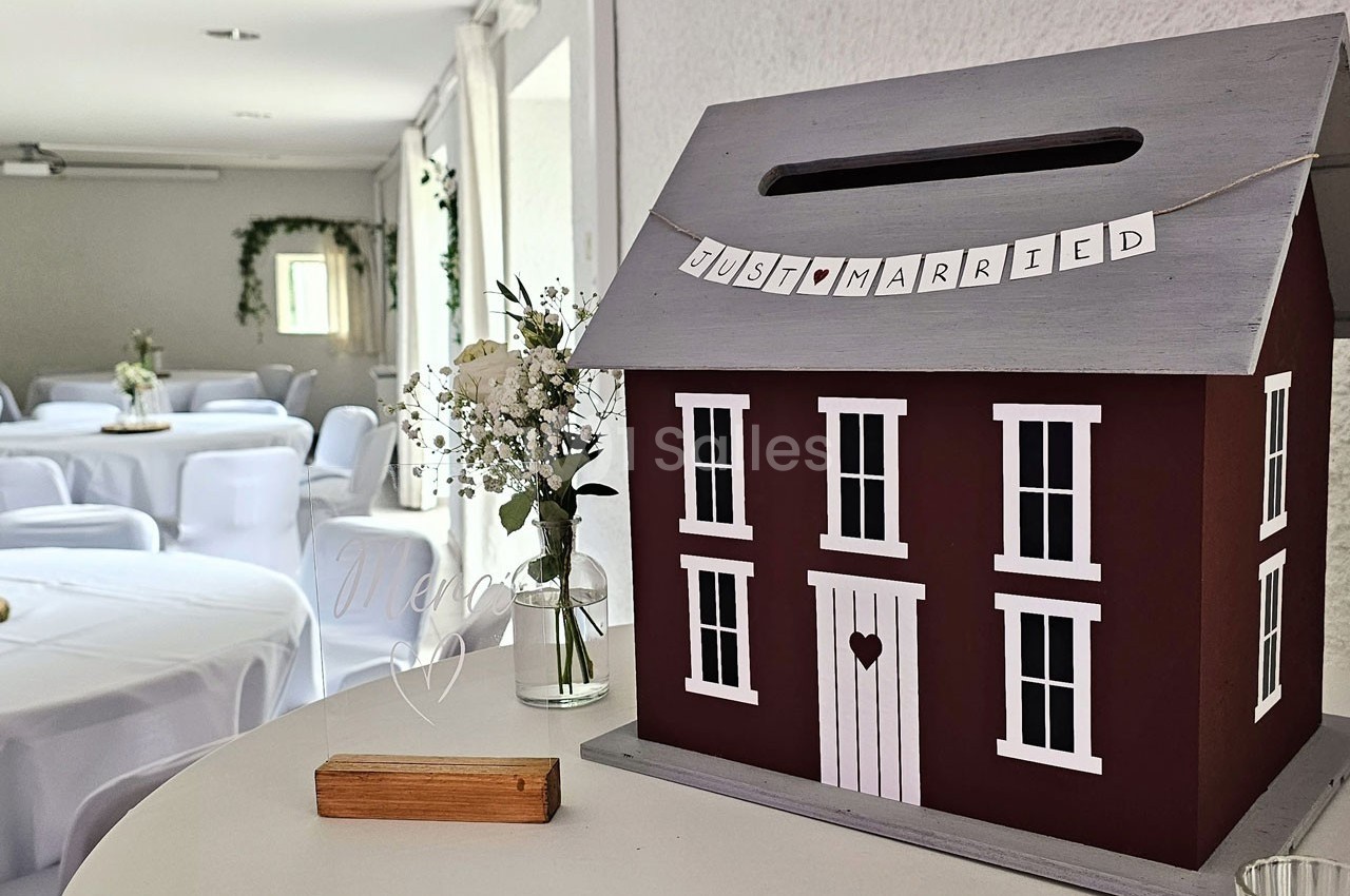 Boîte en forme de maison rouge avec une fente, décorée d'une bannière ’Just Married’, dans une salle de réception.