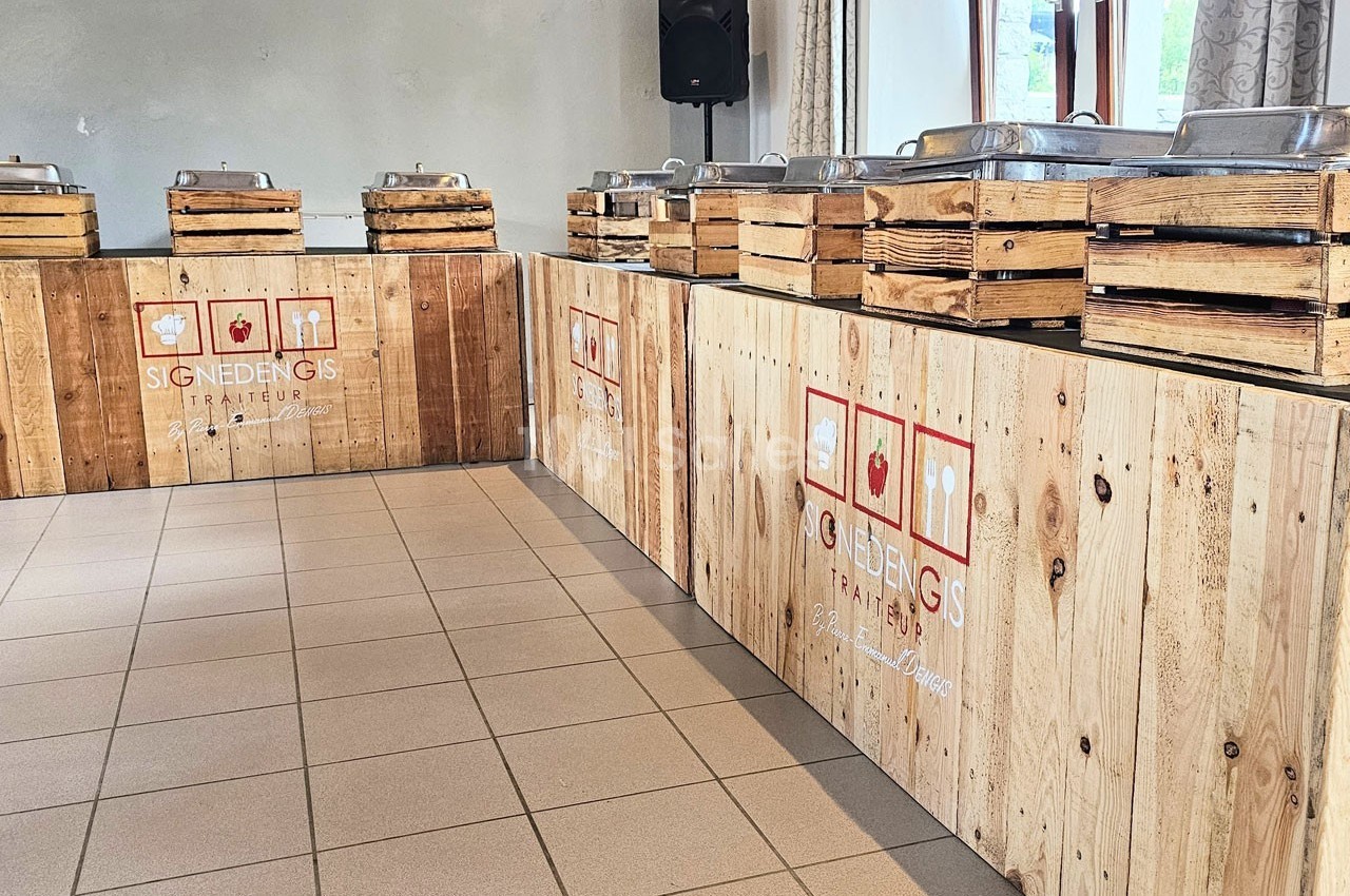 Buffet en bois avec des plats en inox disposés sur des caissons, dans une salle lumineuse avec carrelage au sol.