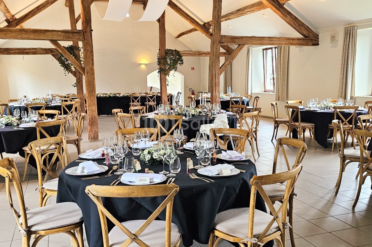 Salle de réception lumineuse avec tables rondes dressées, nappes noires et chaises en bois, décorée pour un événement.