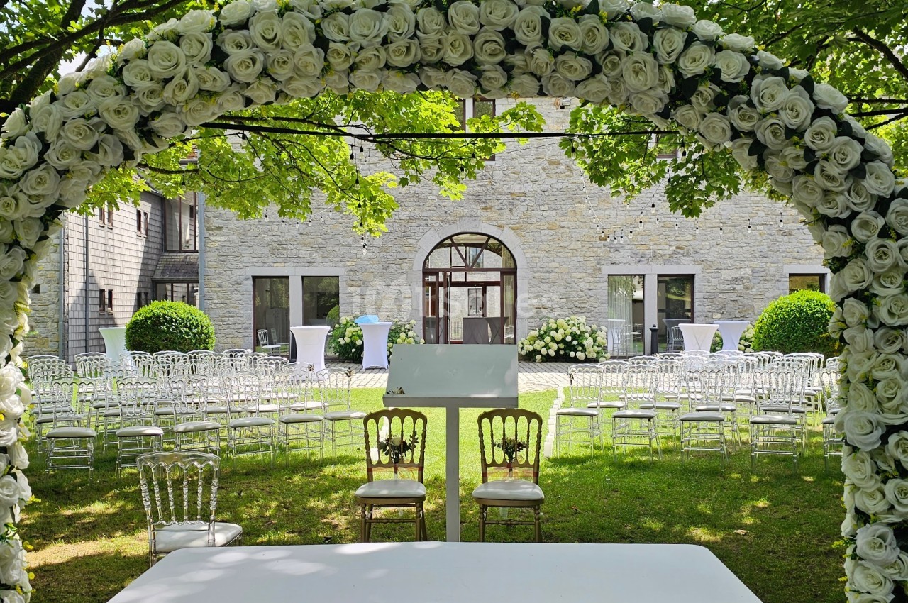 Arche de roses blanches devant des chaises disposées pour une cérémonie en plein air, avec un bâtiment en pierre en arrière…