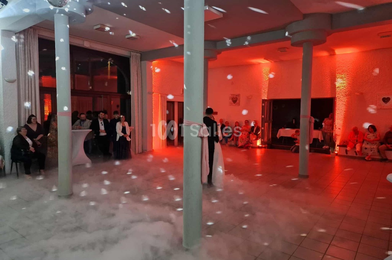 Un couple danse dans une salle éclairée de lumières rouges, entourée de fumée et de quelques invités assis.