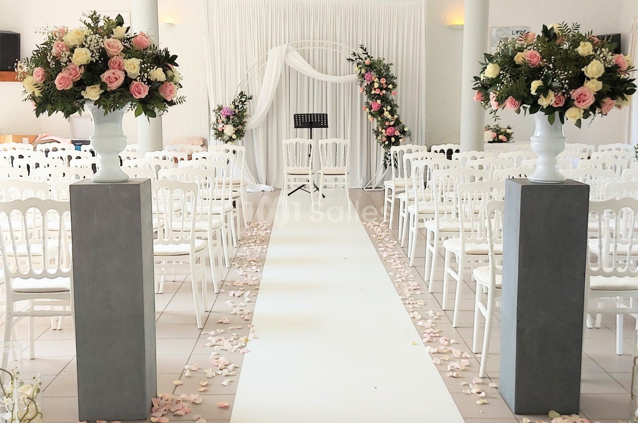 Allée centrale décorée de pétales de rose, entourée de chaises blanches et de compositions florales, menant à une arche…