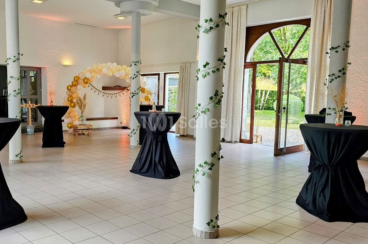 Salle lumineuse décorée pour un événement, avec tables hautes noires, colonnes ornées de lierre et arche de ballons dorés.