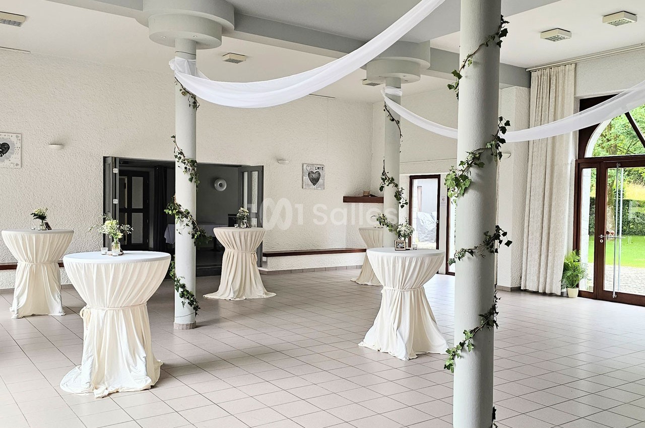 Salle lumineuse décorée de colonnes ornées de lierre et de tables hautes drapées de blanc, avec vue sur un jardin.