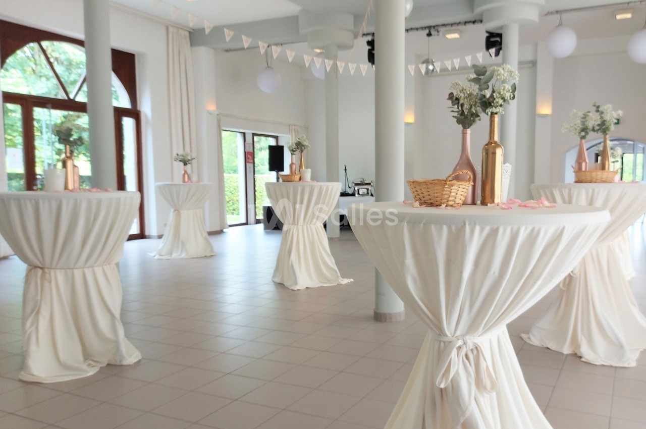 Salle lumineuse décorée pour un événement, avec des tables hautes drapées de blanc et des vases fleuris.