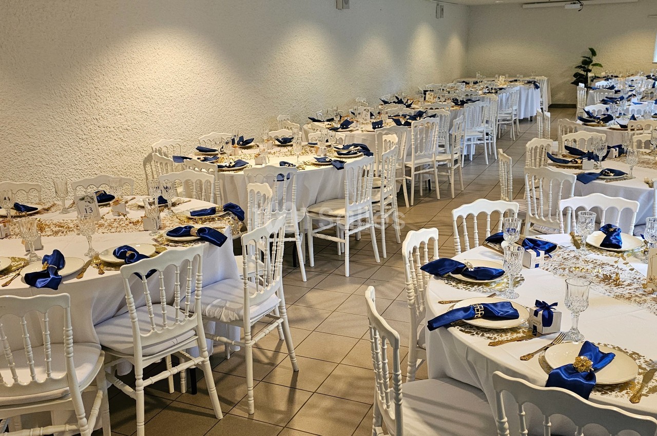 Salle de réception décorée avec des tables rondes dressées, nappes blanches, serviettes bleues et chaises blanches.