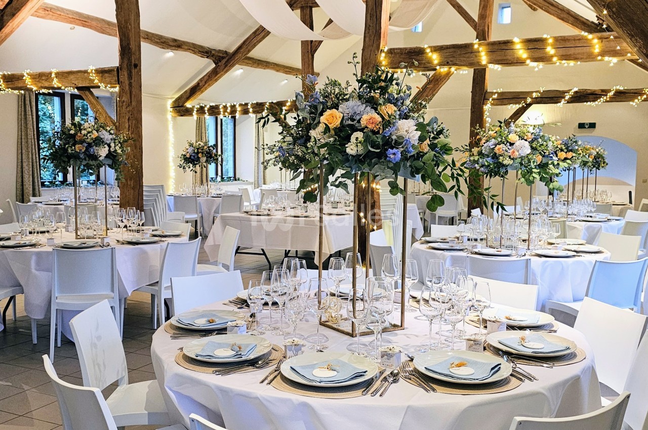 Salle de réception décorée avec des tables rondes, nappes blanches, vaisselle élégante et centres de table fleuris.