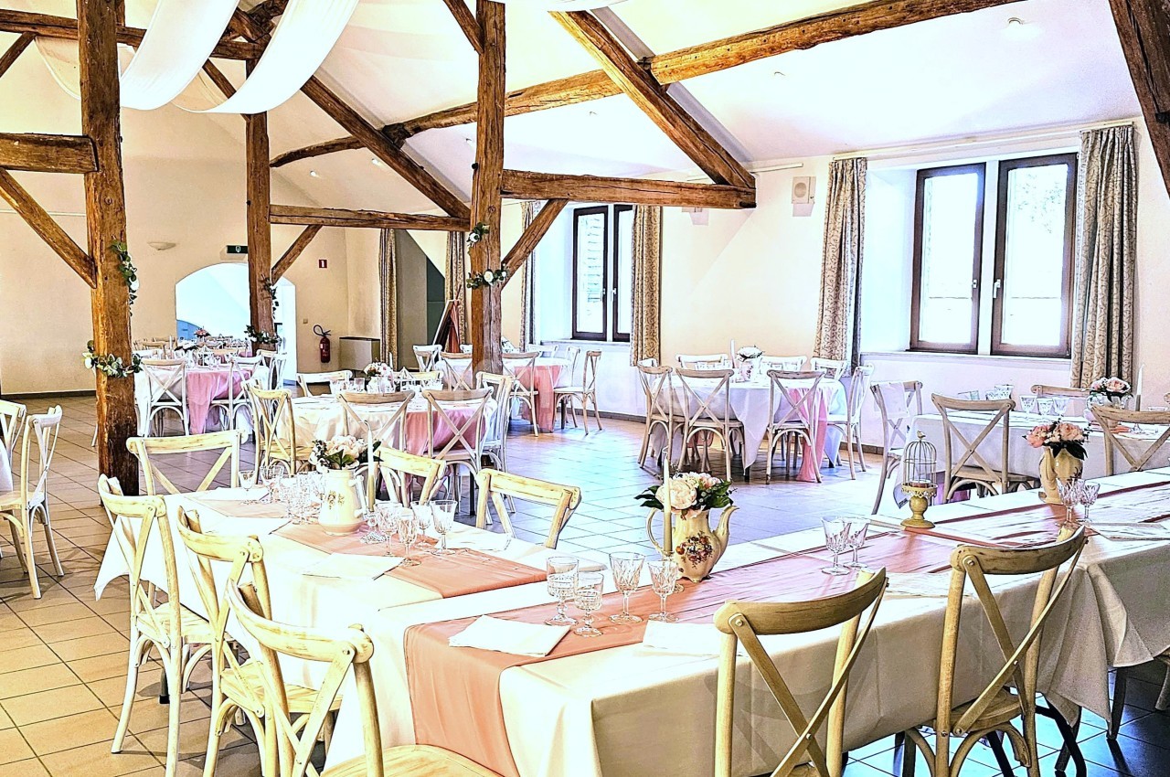 Salle de réception lumineuse avec poutres apparentes, tables décorées de nappes blanches et chemins de table roses.