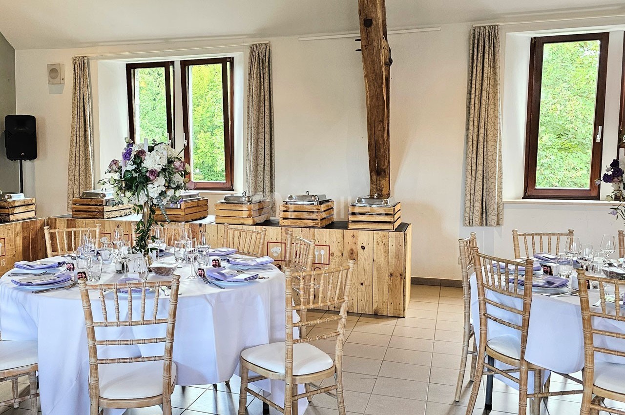 Salle de réception lumineuse avec tables rondes dressées, chaises en bois clair et décorations florales élégantes.