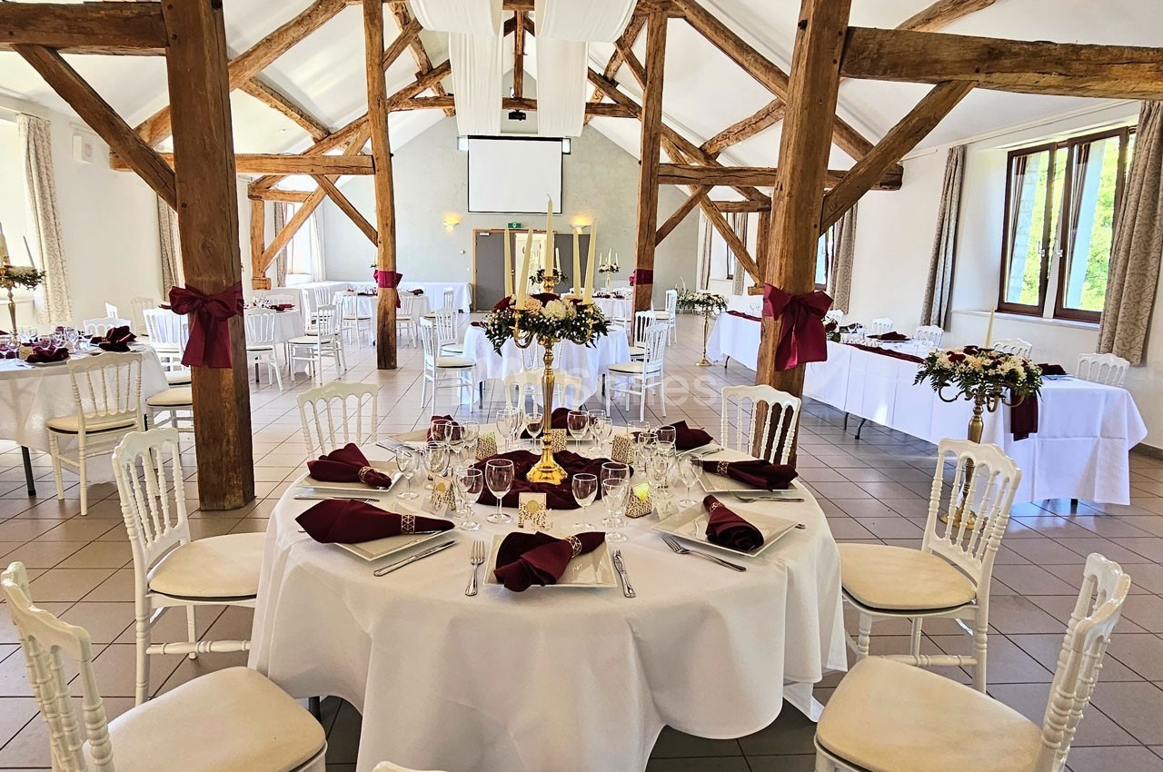 Salle de réception lumineuse avec poutres apparentes, tables rondes dressées et décorations bordeaux et blanches.