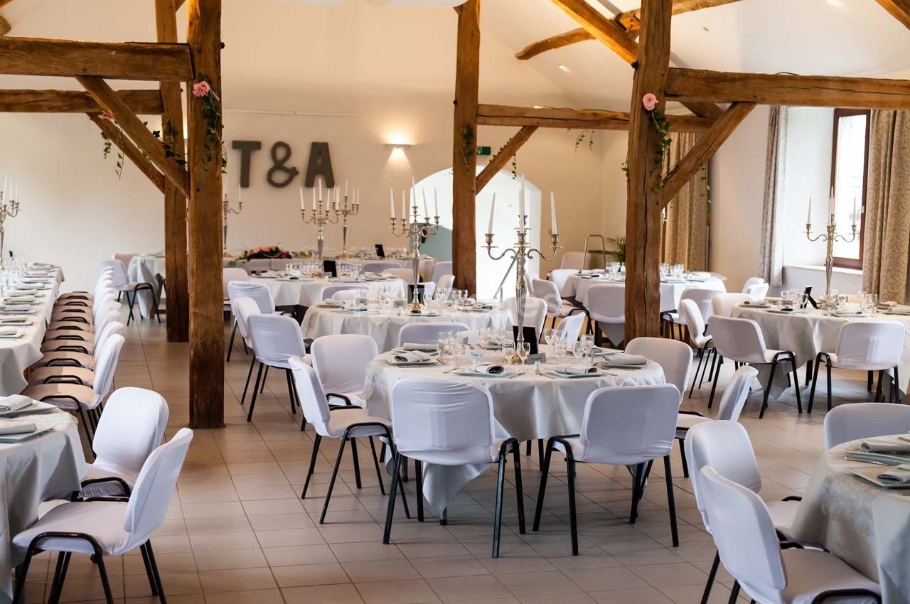 Salle de réception décorée avec des tables rondes dressées, chaises blanches et poutres apparentes en bois.
