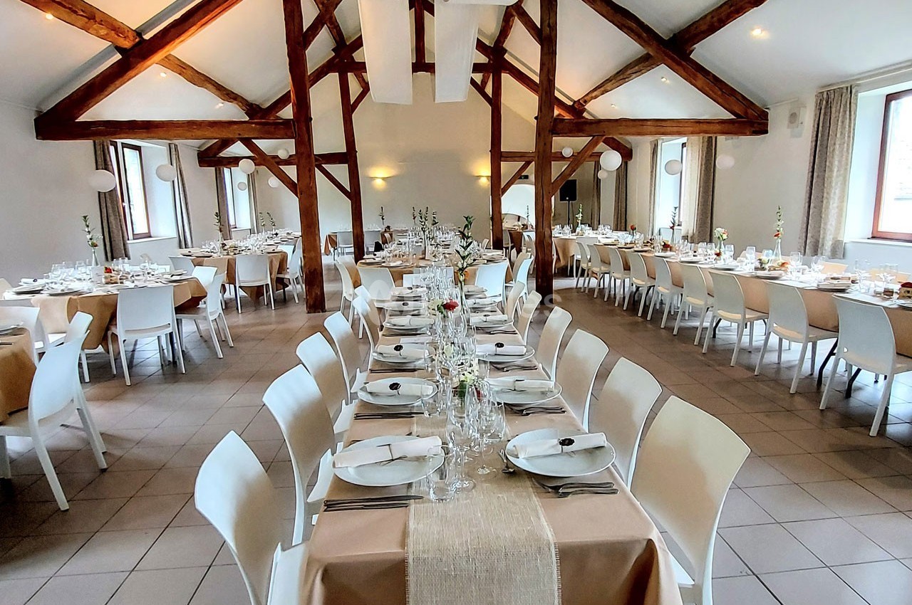 Salle de réception lumineuse avec poutres apparentes, tables dressées pour un repas, nappes beige et décoration élégante.