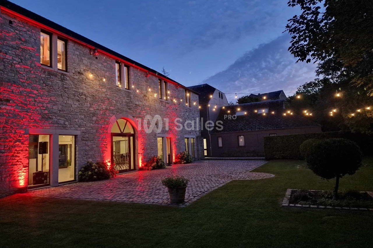 Façade en pierre éclairée par des lumières rouges et guirlandes lumineuses, avec une cour pavée et un jardin au crépuscule.