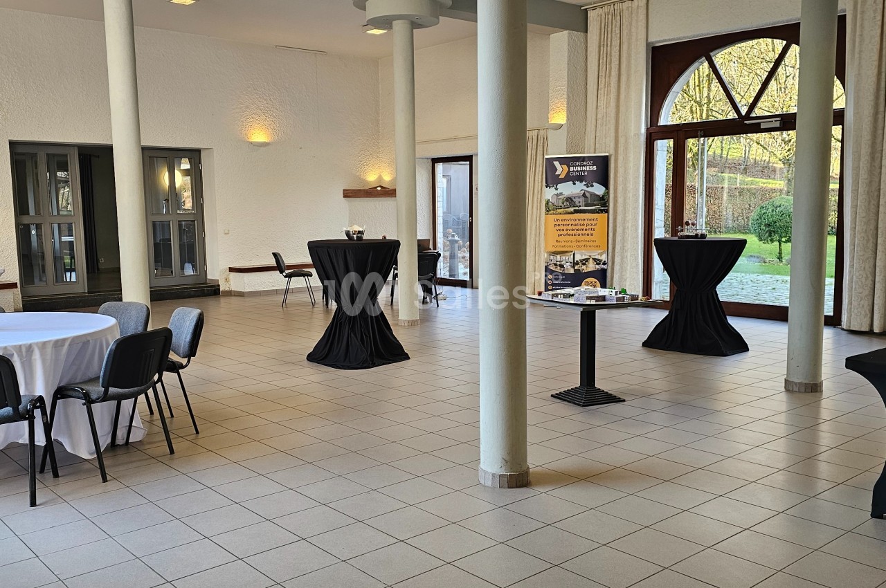 Salle lumineuse avec tables rondes et hautes, nappées de noir et blanc, donnant sur un jardin par de grandes baies vitrées.