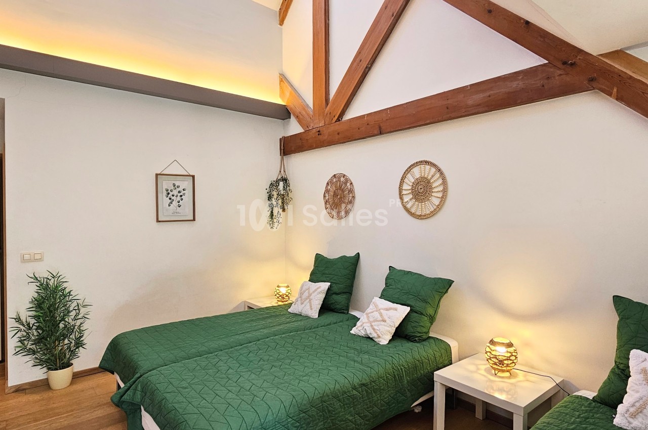 Chambre lumineuse avec deux lits simples, murs blancs, poutres apparentes et décoration végétale et murale.