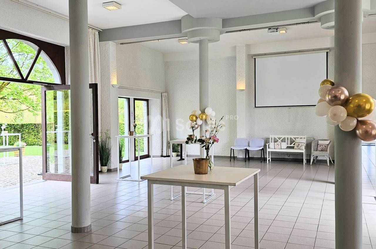 Salle lumineuse avec de grandes fenêtres, décorée de ballons et de fleurs, équipée de tables blanches et d'un écran.