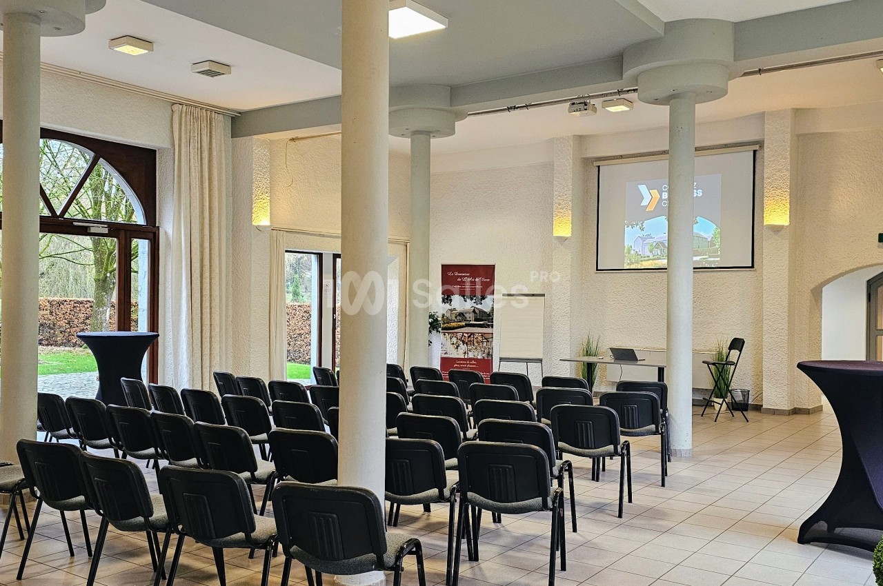 Salle lumineuse aménagée pour une présentation, avec des rangées de chaises, un écran de projection et des tables hautes.