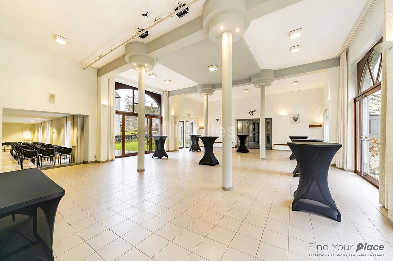 Salle lumineuse avec grandes baies vitrées, tables hautes noires, carrelage clair et vue sur un espace extérieur verdoyant.
