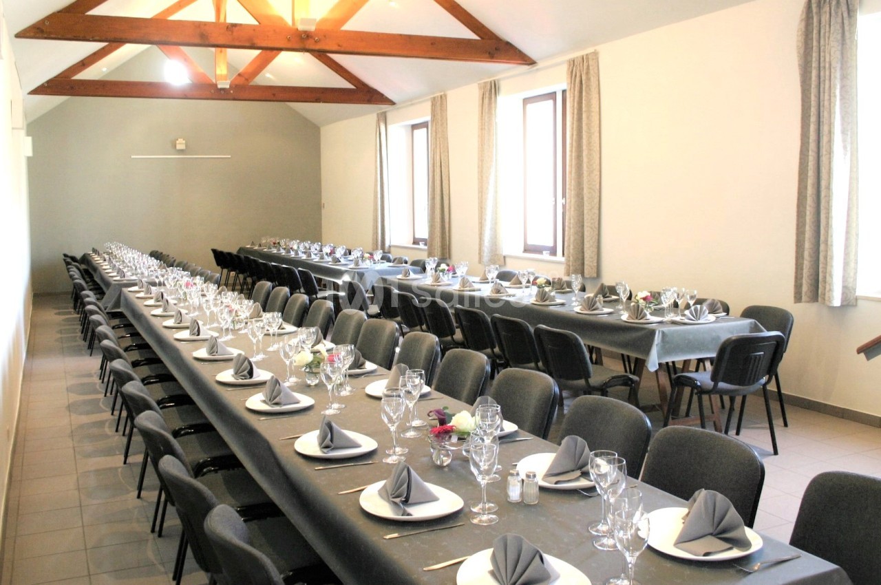 Salle de réception avec deux longues tables dressées, nappes grises, serviettes pliées et verrerie, éclairée par des…