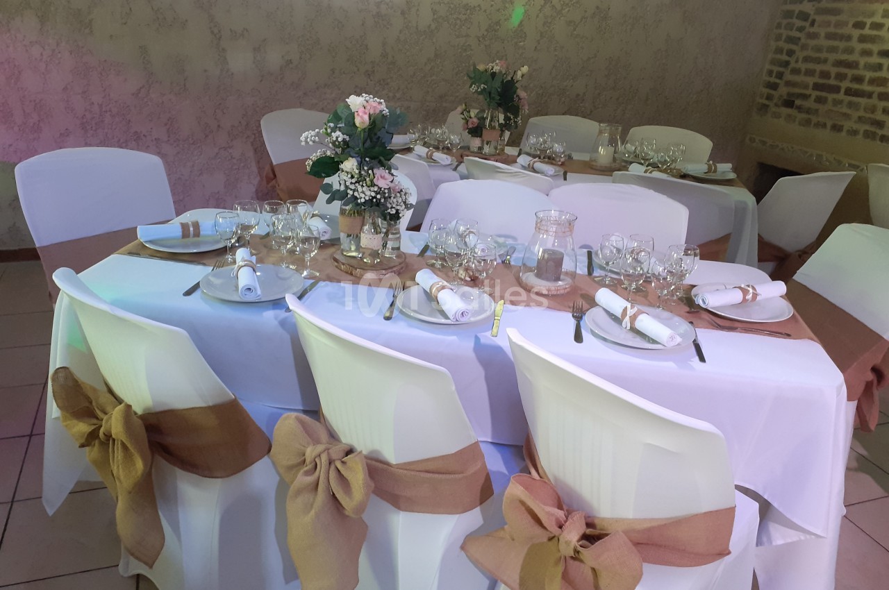 Salle décorée pour un événement avec tables rondes dressées, nappes blanches, rubans beige et compositions florales.
