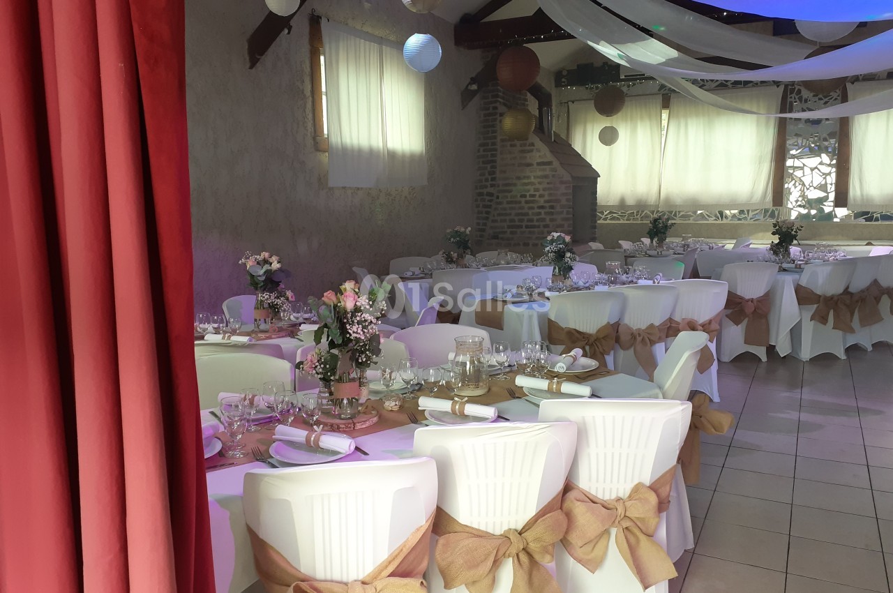 Salle décorée pour un événement, avec tables dressées, chaises ornées de nœuds en toile de jute et éclairage doux.
