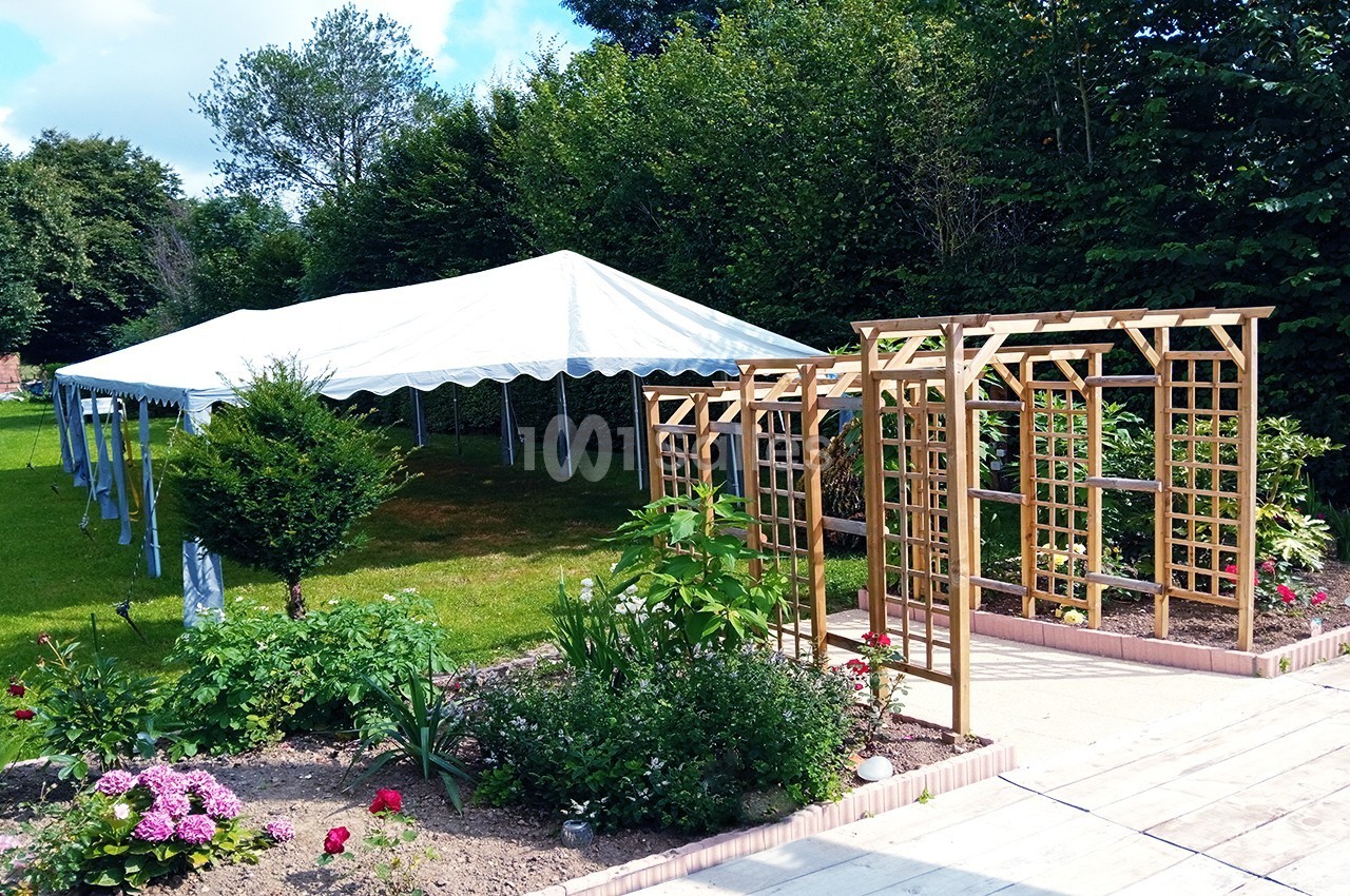 Tente blanche montée sur une pelouse près d'une pergola en bois et d'un jardin fleuri, entourée de verdure.