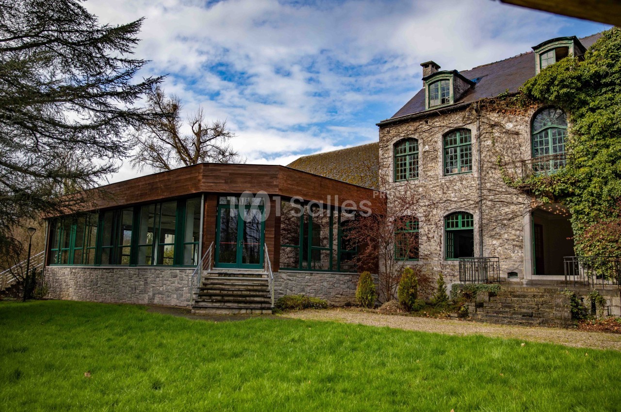 Bâtiment en pierre avec une extension moderne vitrée, entouré de pelouse et d'arbres sous un ciel partiellement nuageux.