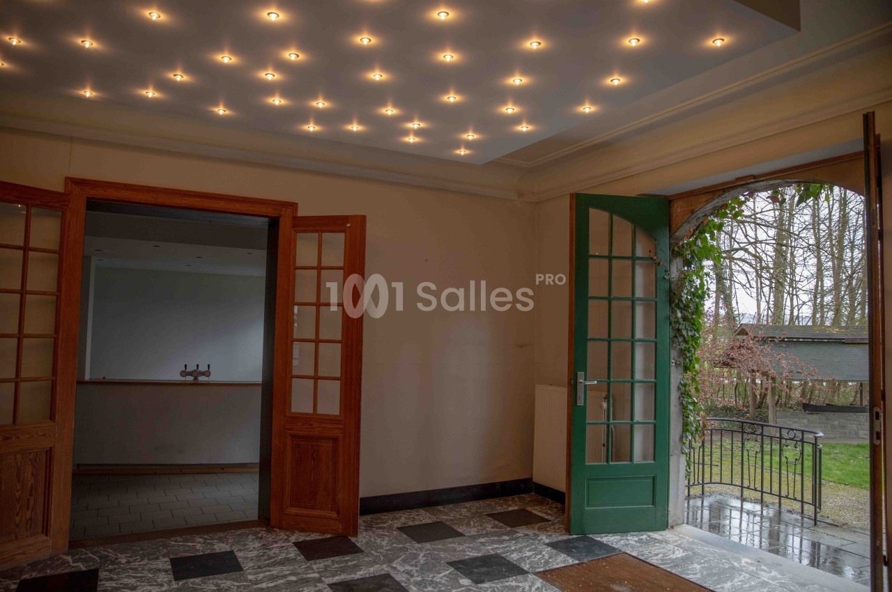 Intérieur d'une pièce avec sol en marbre, portes vitrées en bois, plafond éclairé et vue sur un jardin par une porte ouverte.