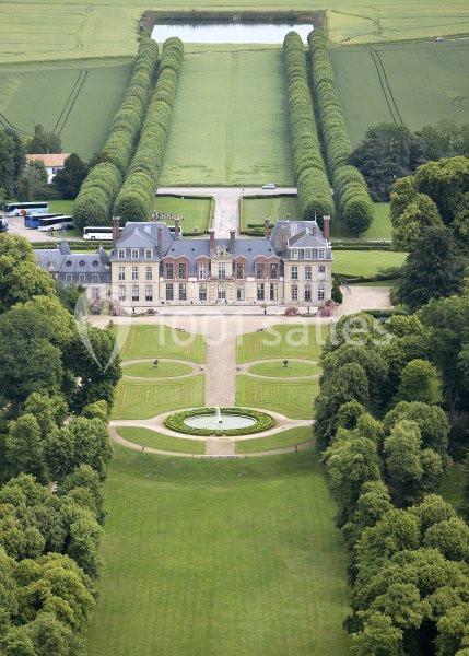 Vue aérienne d'un château entouré de jardins symétriques, d'allées arborées et de champs verdoyants.