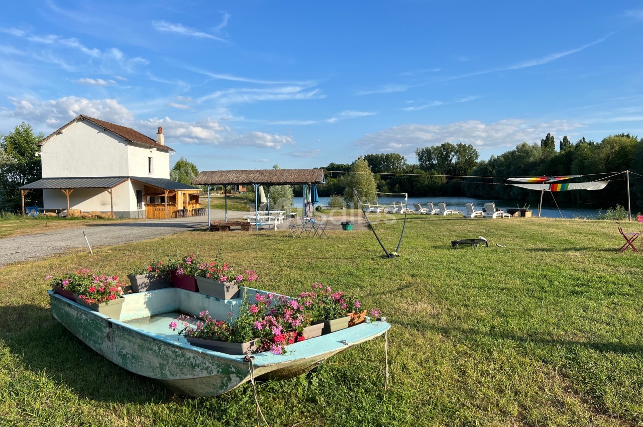 Une barque fleurie sur une pelouse, avec une maison, des tables et un cours d'eau en arrière-plan sous un ciel bleu.