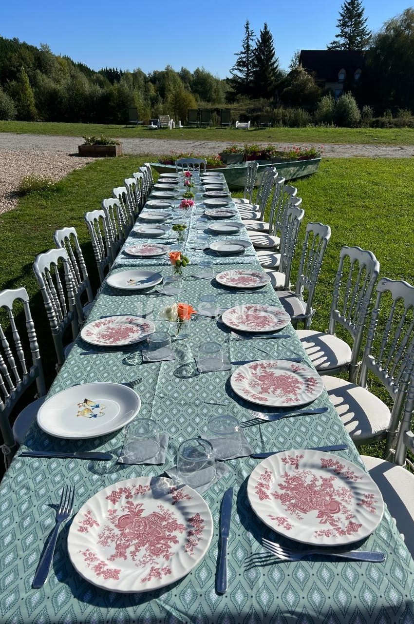 Grande table extérieure dressée avec une nappe verte, des assiettes à motifs rouges et des chaises blanches, dans un cadre…
