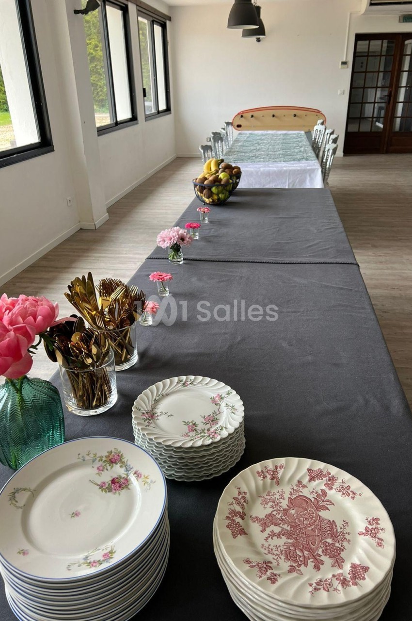 Table dressée avec une nappe noire, vaisselle empilée, couverts dorés, fleurs et corbeille de fruits dans une salle…