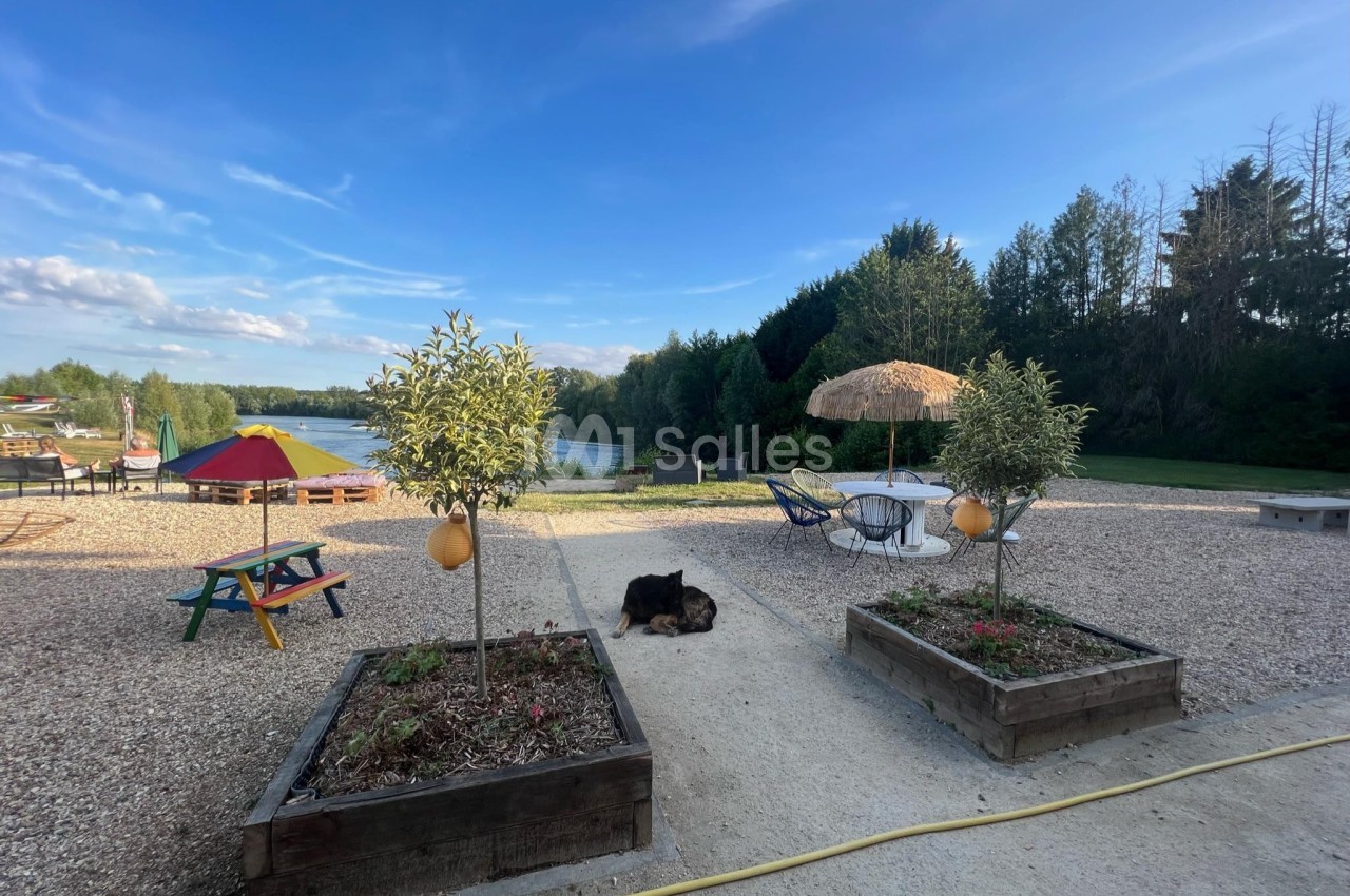 Espace extérieur avec tables, parasol en paille, arbres en pot, un chien couché et vue sur un plan d'eau entouré de verdure.