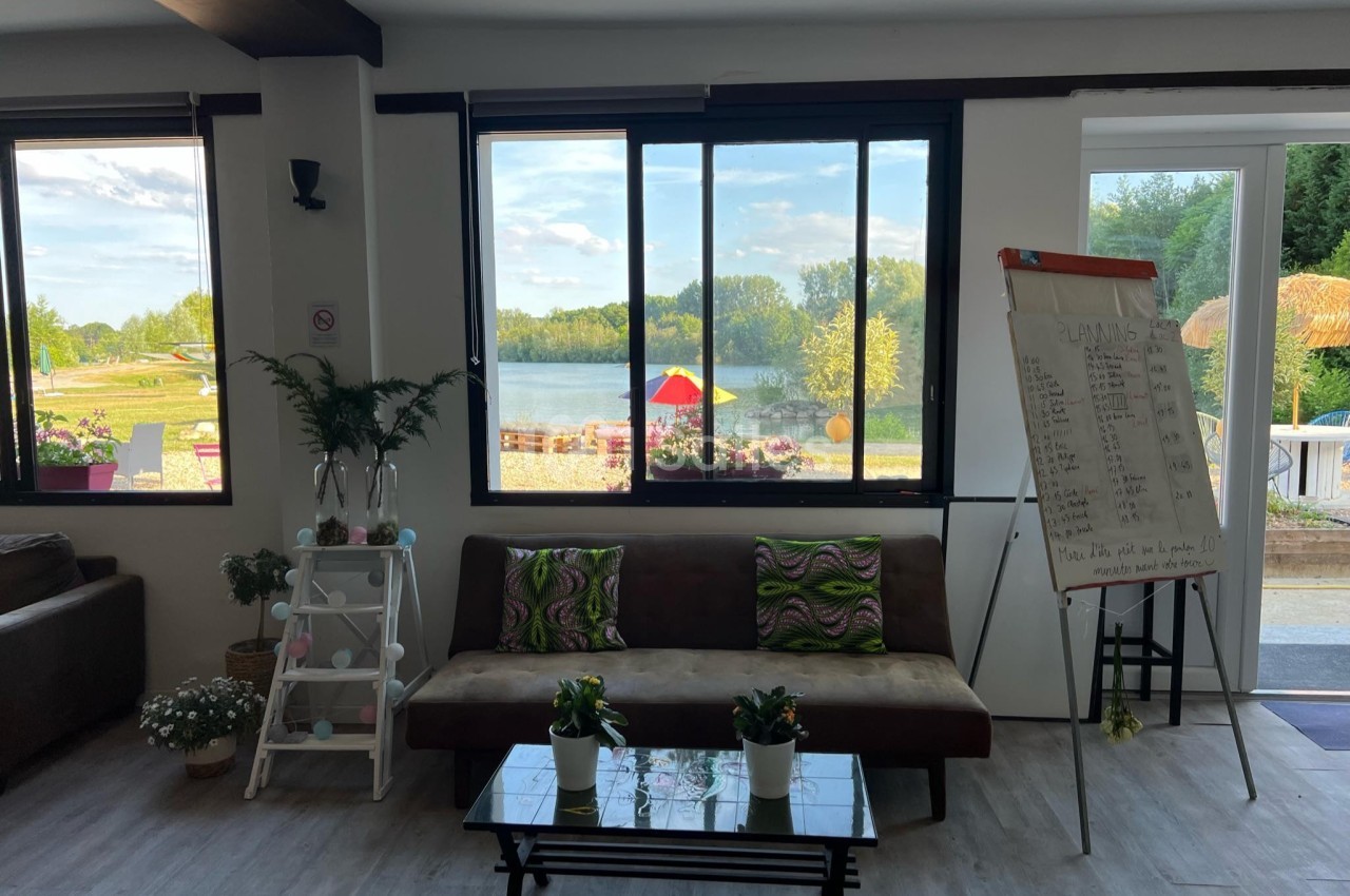 Salon lumineux avec canapé, table basse, plantes, tableau de notes et vue sur un lac à travers de grandes fenêtres.