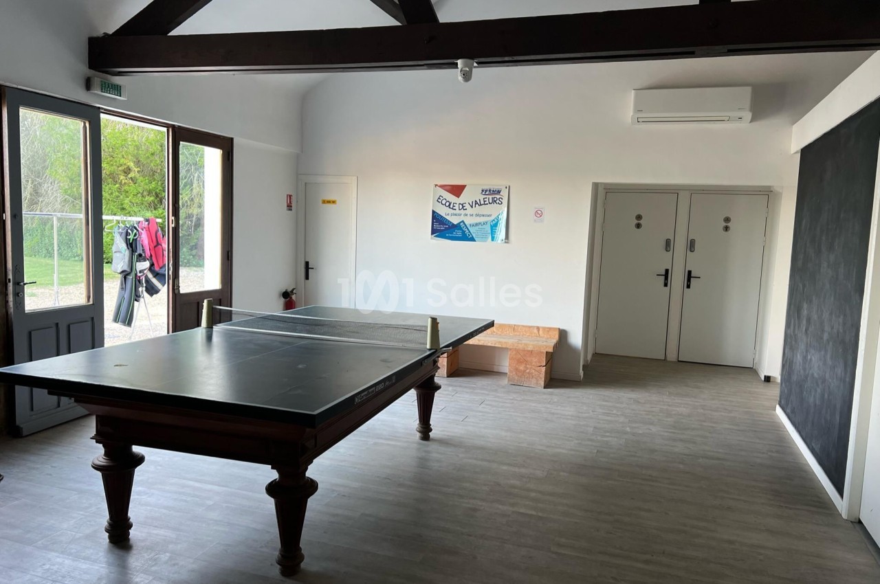 Salle lumineuse avec une table de ping-pong au centre, des portes fermées et un tableau noir sur le mur droit.