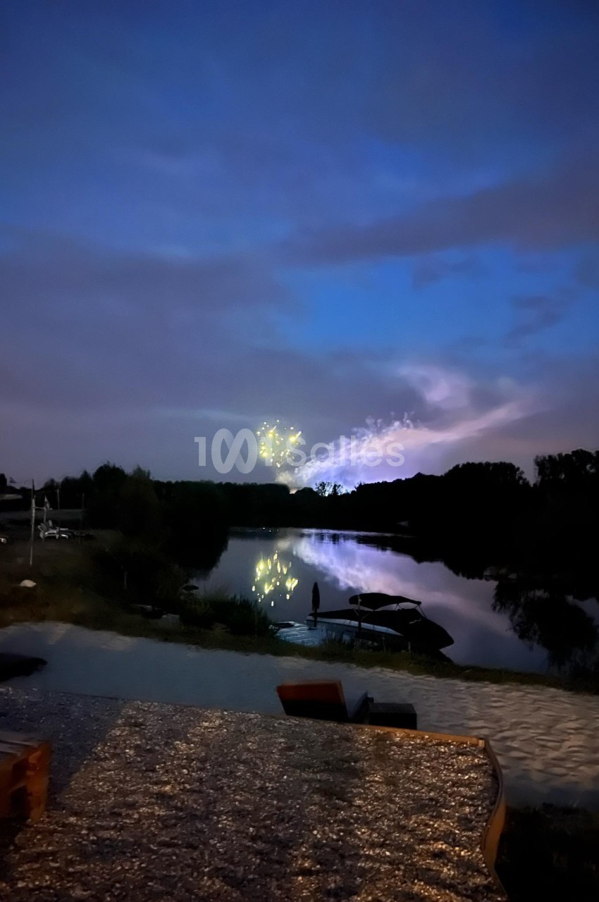Ciel nocturne avec des feux d'artifice reflétés sur un lac calme, entouré de végétation et d'un rivage éclairé.