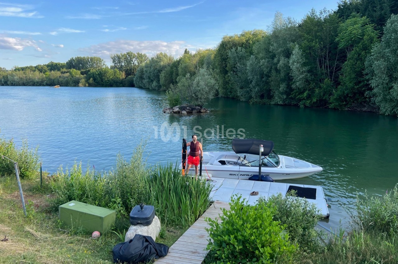 Un homme debout sur un ponton en bois près d'un bateau amarré, entouré d'un lac et de végétation.