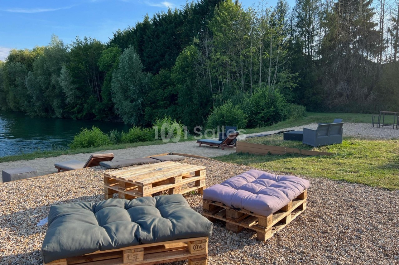 Mobilier en palettes avec coussins installé sur un sol gravillonné, près d'un plan d'eau entouré de végétation.
