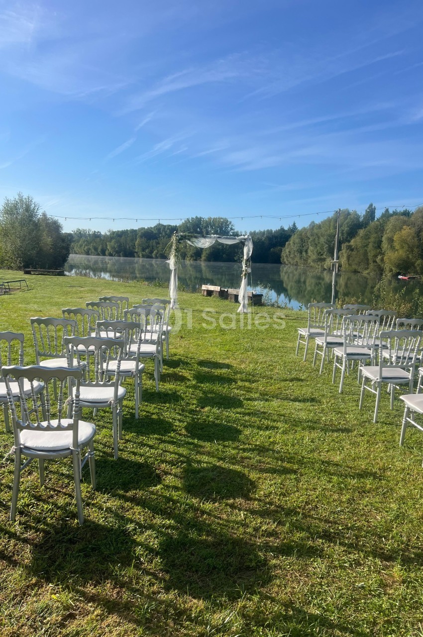 Chaises blanches disposées en extérieur face à une arche décorée, près d'un lac sous un ciel bleu.