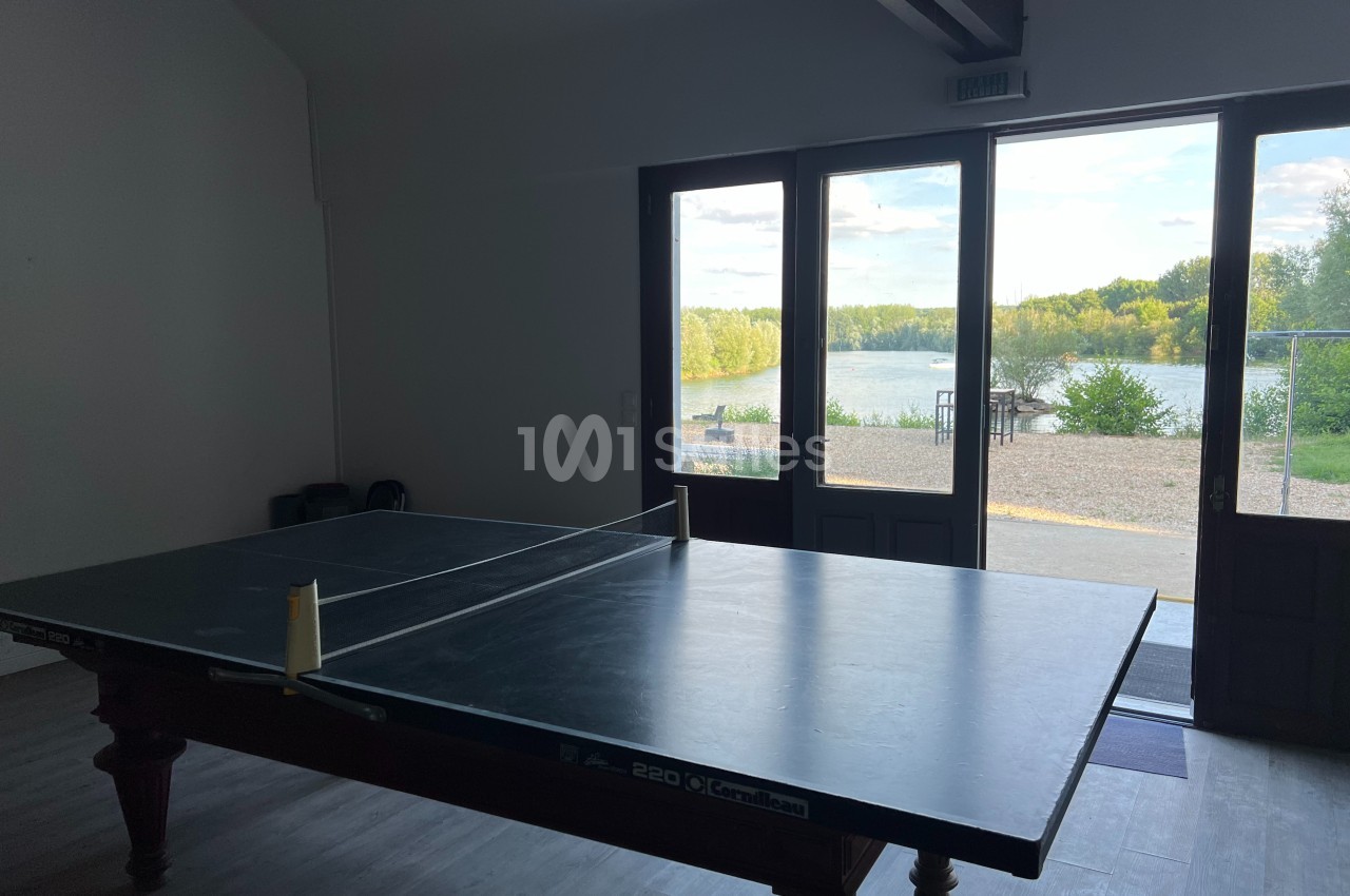 Table de ping-pong dans une salle sombre avec vue sur un lac et des arbres à travers des portes vitrées.