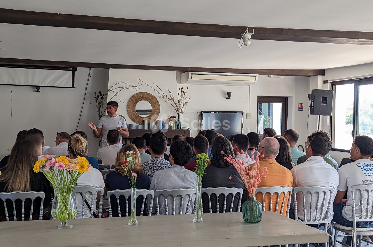 Un groupe de personnes assises écoute un intervenant dans une salle lumineuse avec des décorations florales.