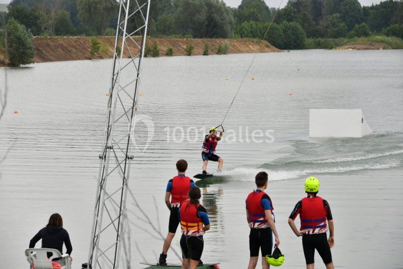 Un groupe de personnes en gilets de sauvetage observe un wakeboarder sur un lac équipé d'un système de téléski nautique.