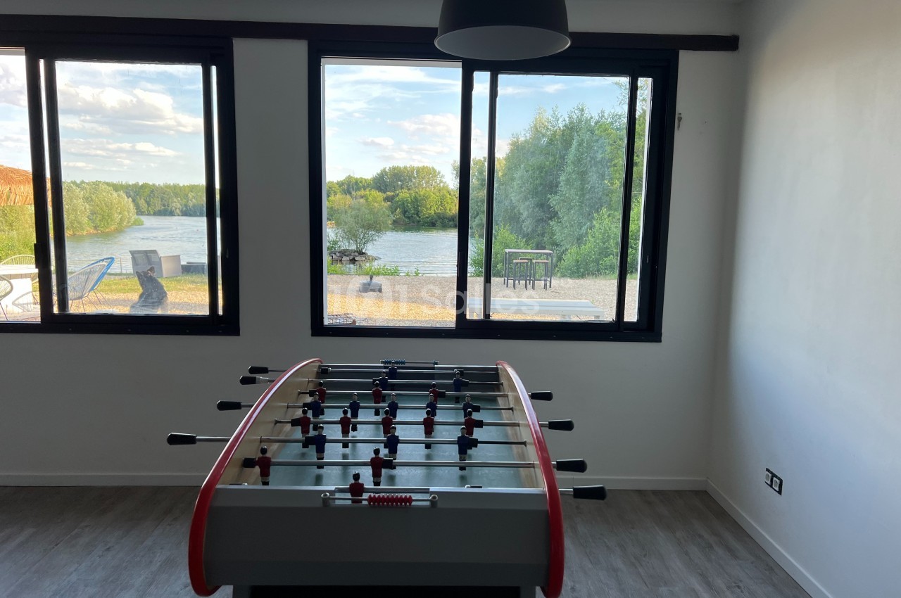 Table de baby-foot dans une pièce lumineuse avec vue sur un lac et des arbres à travers de grandes fenêtres.