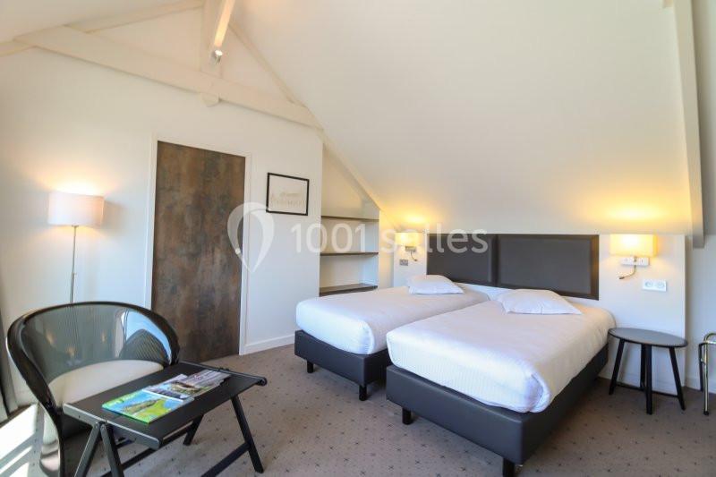 Chambre lumineuse sous combles avec deux lits simples, un fauteuil, une table basse et une porte en bois.