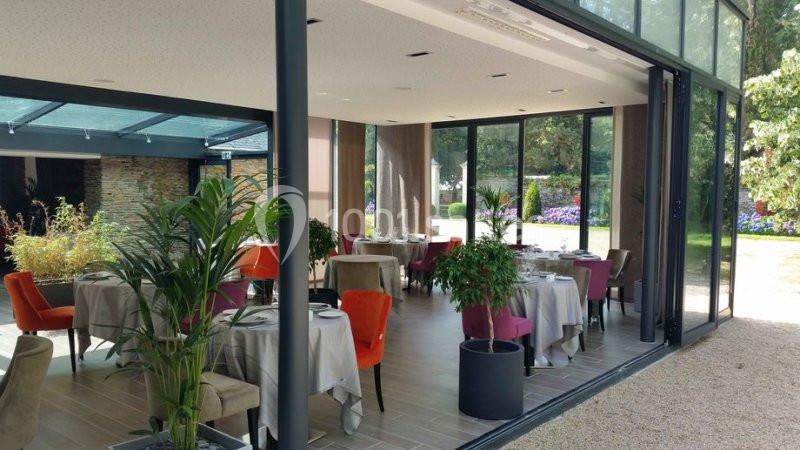 Salle de restaurant lumineuse avec baies vitrées ouvertes sur un jardin, tables dressées et chaises colorées.
