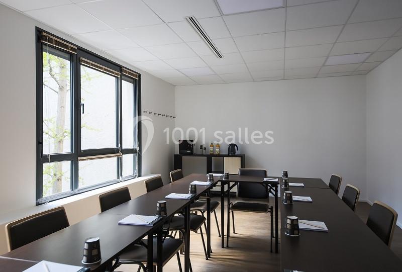 Salle de réunion lumineuse avec tables en U, chaises, blocs-notes, gobelets et coin boissons au fond.